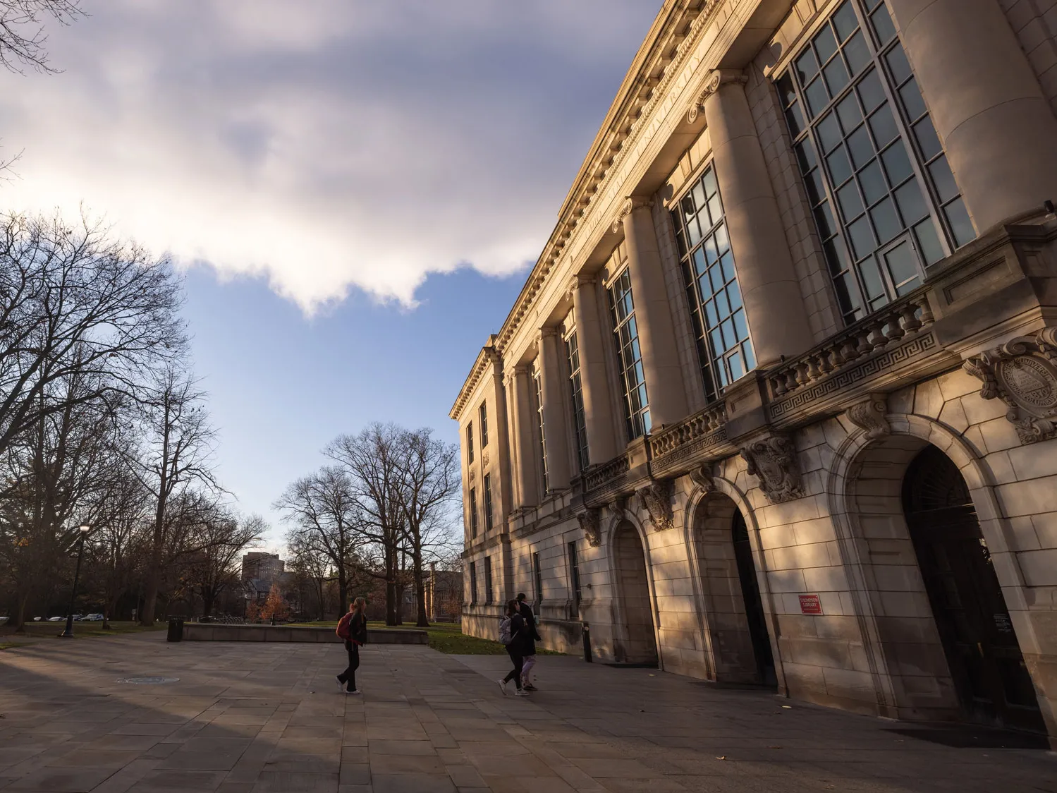 In the morning light on a cold day (indicated by leaf-less trees and the distant people walking toward the library while wearing winter coats), clouds against a blue sky cast shadows on the columned front of the library. Multi-paned windows reflect the blue of the sky above a few arched entrances. 