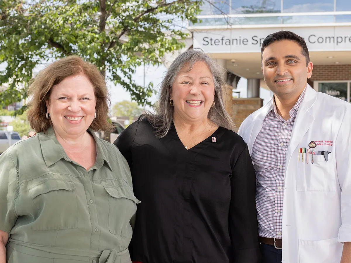 Three people pose for this photo in front of the entrance to the Stefanie Spielman Comprehensive Breast Center, part of The Ohio State University Comprehensive Cancer Center – Arthur G. James Cancer Hospital and Richard J. Solove Research Institute. The three are Molly Ranz Calhoun and the doctors who treated her cancer. They stand closely as they pose, seeming like they have a good relationship, and all smile as they squint a bit in the bright sunshine.