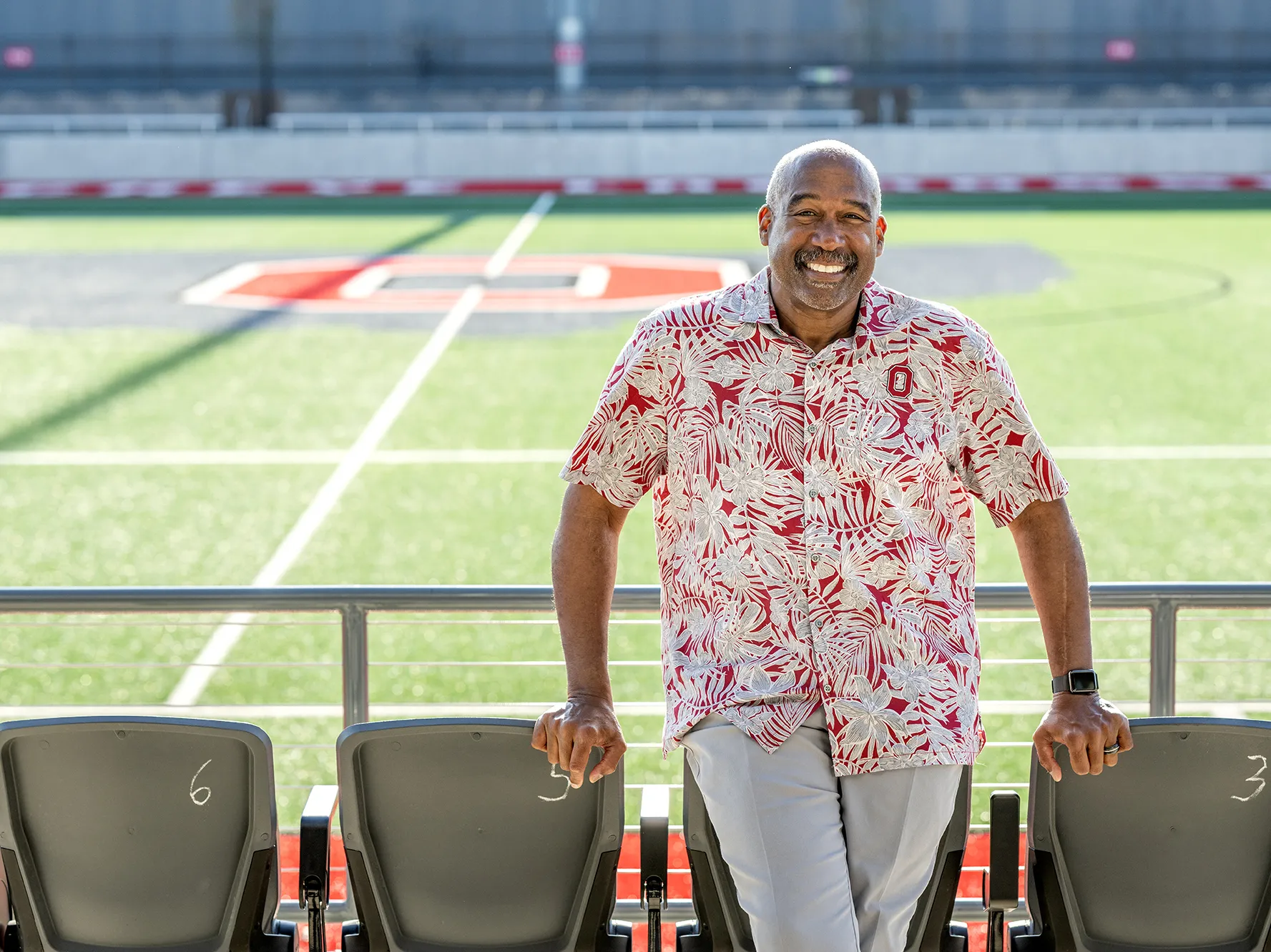 Wearing an Ohio State-colored tropical shirt—that looks perfect for retirement or vacation—Gene Smith grins as he leans on some front-row seats at Ohio Stadium. 