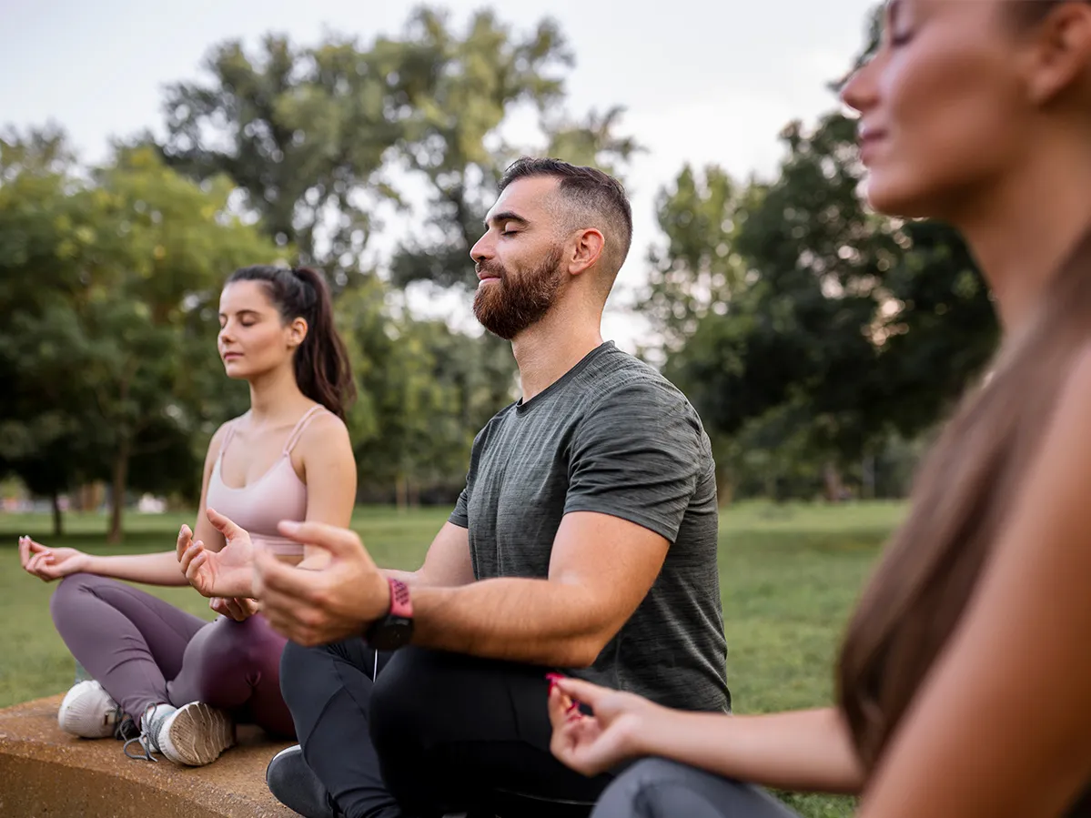 In a stock image, three people sit in a park in a relaxing yoga pose.
