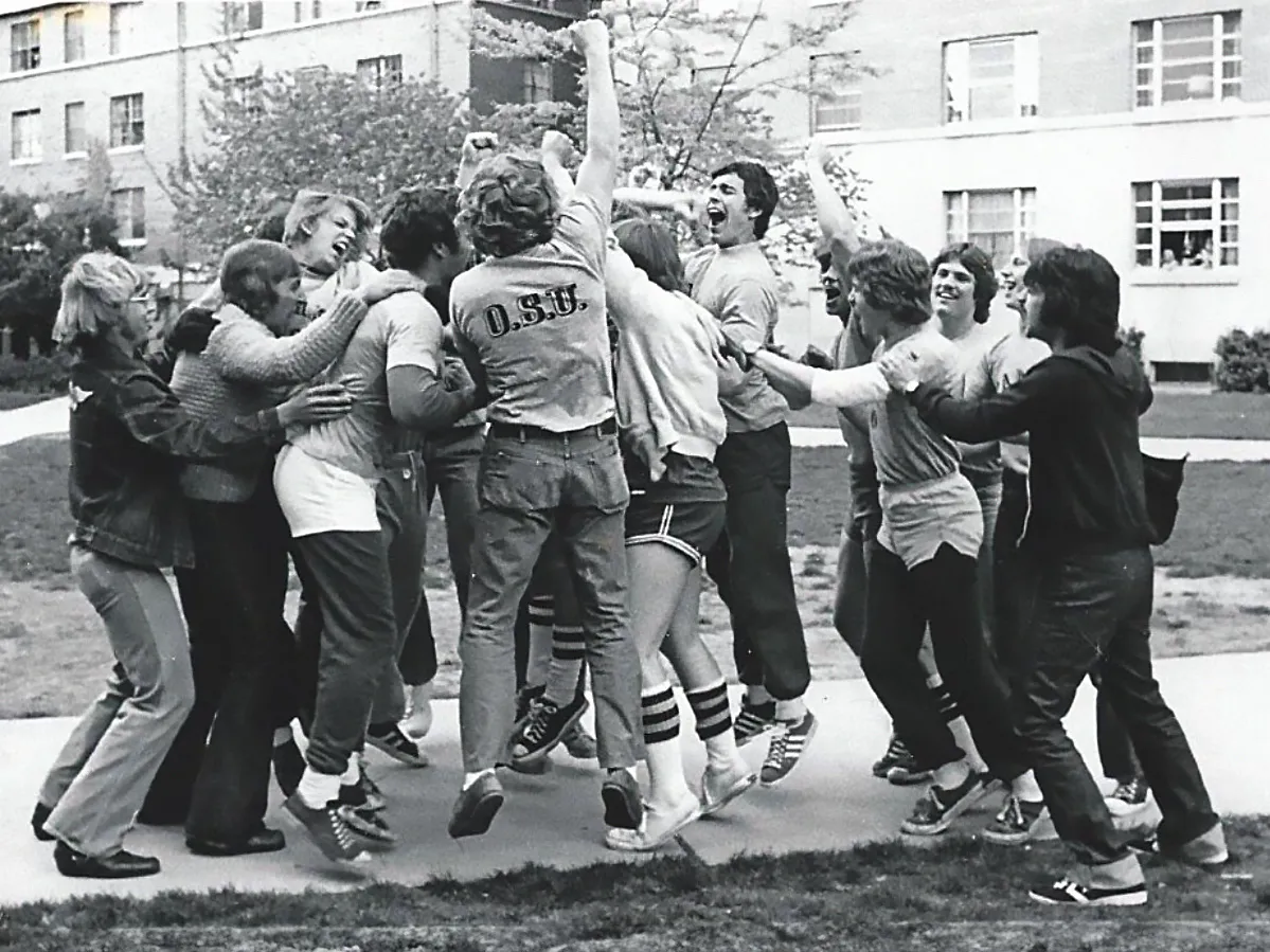 A group of at least ten men, some leaping vertically, unite with arms embracing or thrust in the air in celebration of victory in a tug of war contest.