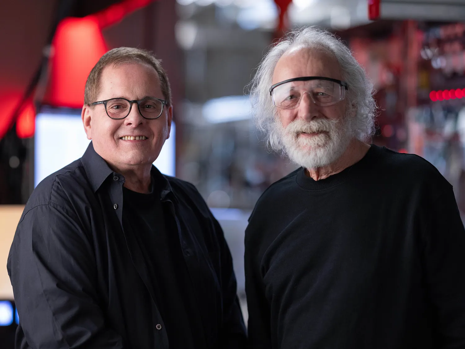 Lou DiMauro and Pierre Agostini smile as they pose for a portrait. Pierre has flowing white hair and a beard and wears safety goggles. Lou has short brown hair and glasses. Both are white men who radiate friendliness while dressed casually. 