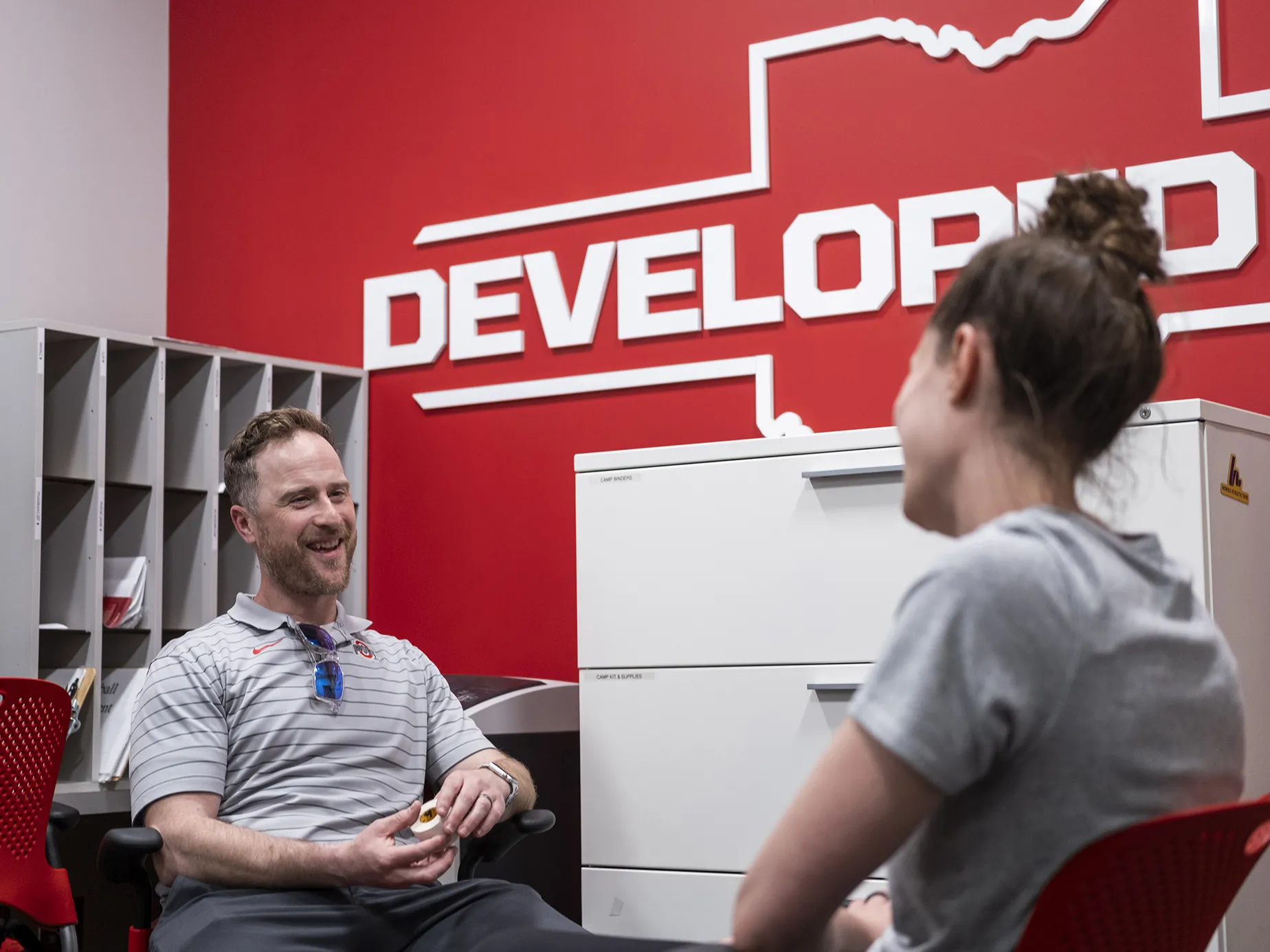 Jamey Houle, a white man in a striped Ohio State golf shirt, laughs as he speaks with a woman shown from the back. They’re both sitting in chairs in an office in the Department of Athletics building. Jamey is a white man with wavy hair and he’s looking directly at his colleague as they chat.