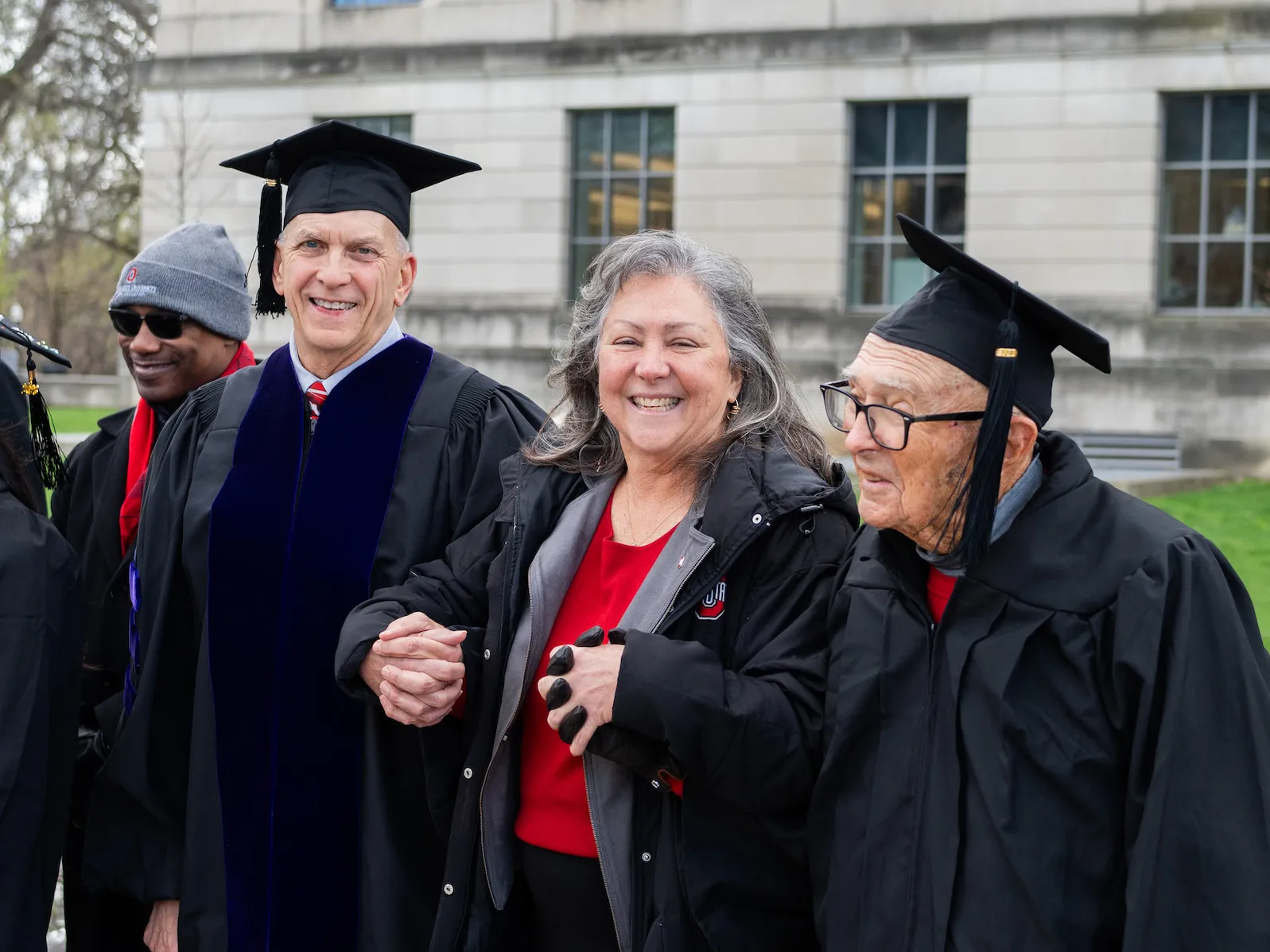 Molly Ranz Calhoun grins happily as she holds hands with the men standing on each side of her—one is her elderly father; the other is a taller man standing up straight and smiling proudly. Both men wear the types of gowns and hats put on for graduation ceremonies. They stand outside on the Oval for a ceremony.