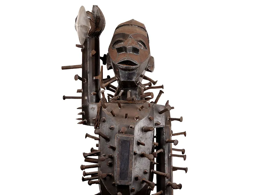 A metal statue with a weathered patina shows an African warrior holding up his sword with thick nails or spikes hammered all over his body. The exception is his face—none is higher than his chin. The warrior looks straight forward. What would usually be rounded—shoulders, belly, arc of the cheek, is rendered in flat panels of metal, adding a geometric look to the warrior. 