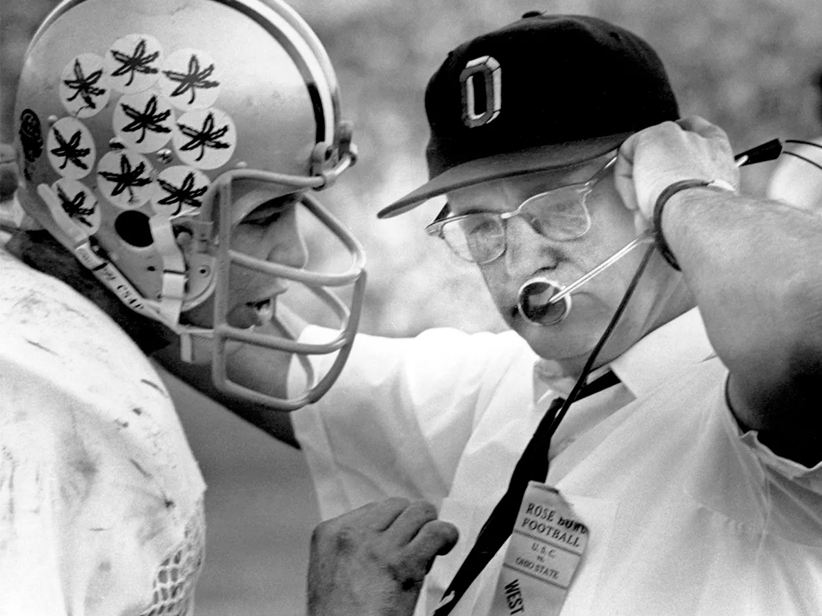 Legendary football coach Woody Hayes stands with Ohio State quarterback Rex Kern, with his arm on his shoulder, as he talks into his headset to players on the field. The image is from Ohio State's archives, and Rex has the trademark buckeye leaf stickers on his helmet, each awarded for an outstanding play.