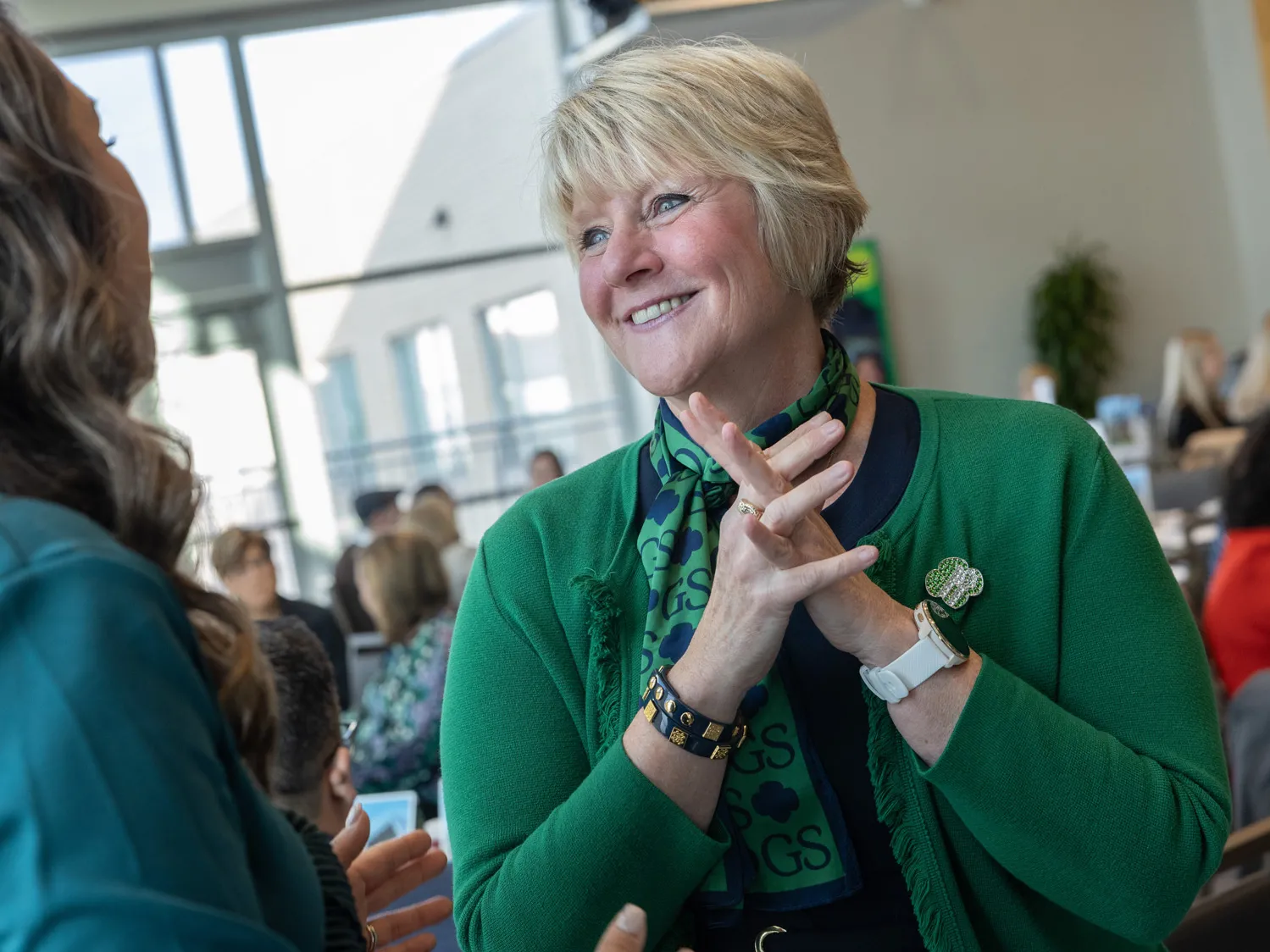 From this photo, you can tell that Tammy Wharton, a white woman with short blond hair and a pretty smile, has excellent listening habits. She seems super-engaged and even encouraging as she looks directly at the woman who’s speaking with her, who is pictured from behind. Other people are in the background of the photo, so you can see it’s a meeting or convention at a place where huge windows let in lots of natural light. 