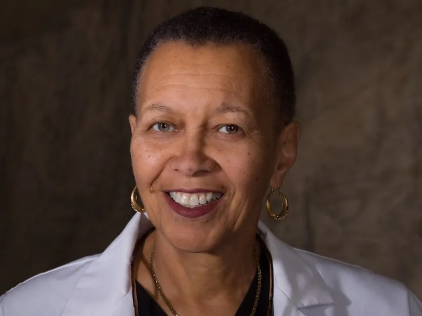 In a portrait showing her head and shoulders, Romaine Bayless smiles while wearing her lab coat, gold hoops earrings and a pair of gold necklaces. A black woman with close cropped hair, a pretty and wide smile, and eyes that crinkle as the corners, her expression says she is friendly and smart.