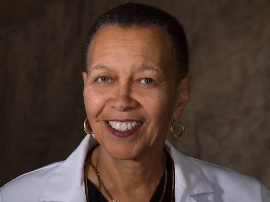 In a portrait showing her head and shoulders, Romaine Bayless smiles while wearing her lab coat, gold hoops earrings and a pair of gold necklaces. A black woman with close cropped hair, a pretty and wide smile, and eyes that crinkle as the corners, her expression says she is friendly and smart.