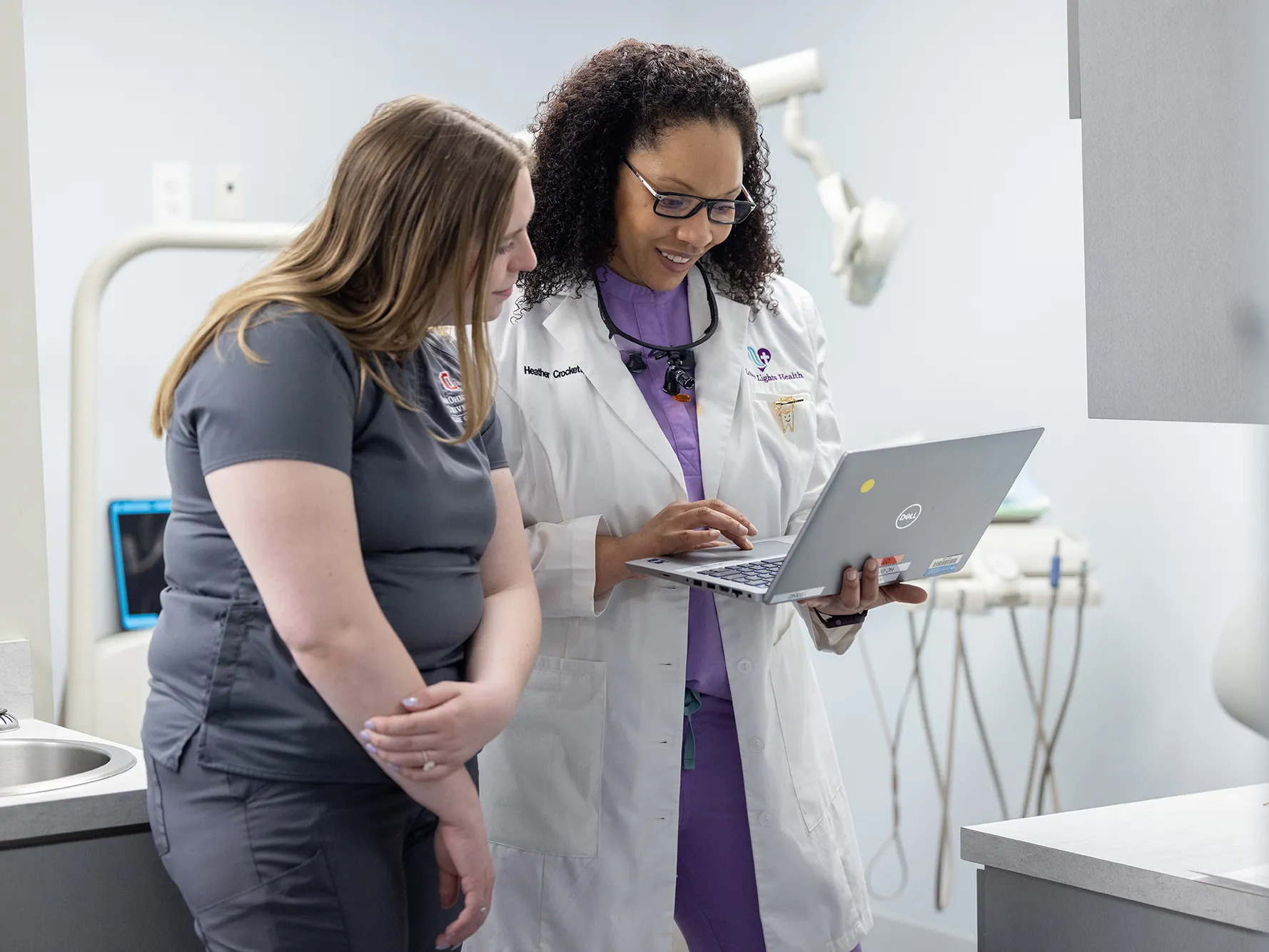 A student in Ohio State scrubs leans in to see the laptop held by a professional dentist wearing a white doctor’s coat. The dentist is a black woman with curly hair and glasses. She’s smiling as she shows the student the screen. The student is a young white woman with long hair and she seems thoughtful.