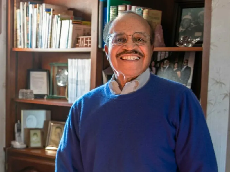 Mac Stewart, a black man with glasses, a genuine smile and a button up under his sweater, poses iin front of a book shelf at his home.