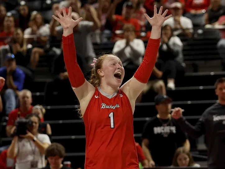 Sarah Morbitzer, a young white woman with her red hair pulled back in a braid, cheers a good play while on the volleyball court. She wears an Ohio State volleyball uniform, with sleeveless shirt, short black shorts, kneepads and arm wraps. The stands and fans are in the background.