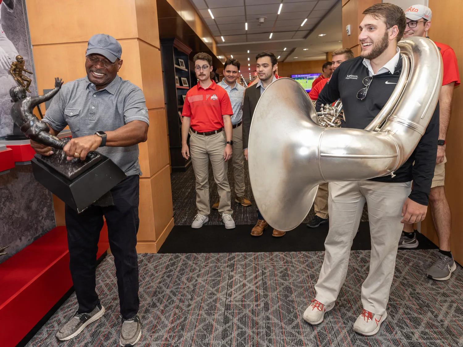 Archie Griffin hefts a replica of his Heisman Trophies—it must be heavy because you can see the muscles in his forearms. Watching are members of the marching band. The student in the front, a young white man with a beard, has a sousaphone wrapped around him. This was the day they invited Griffin to dot the i at a game this season. 