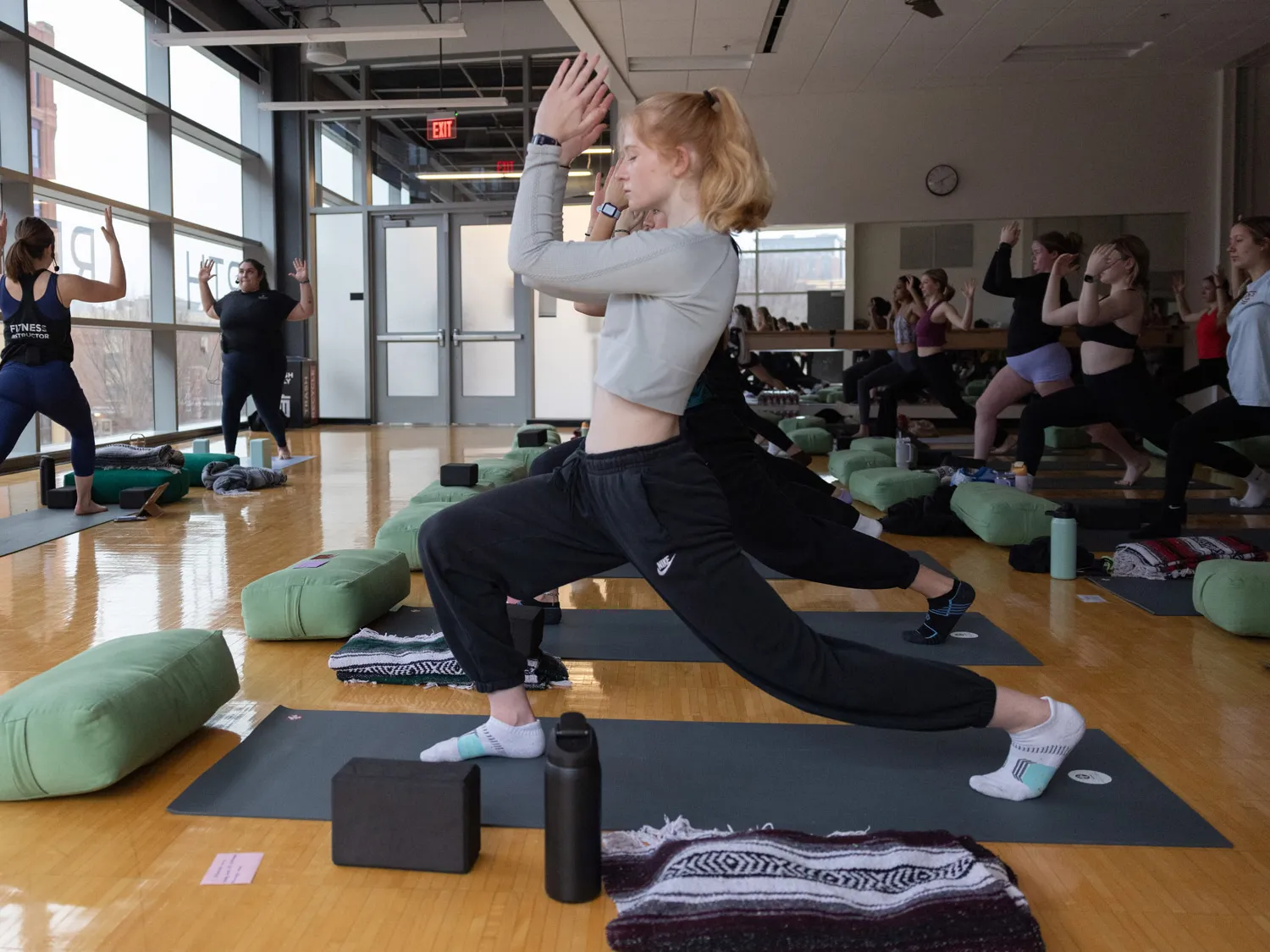 Lines of Buckeye students, each on their own yoga matte, strike the yoga pose shown by the teachers. It is right leg forward and bent so the flat of the foot is on the floor; left leg stretched far behind, so only the ball of the foot is on the floor; and elbows bent 90 degrees and hands held just above head height.