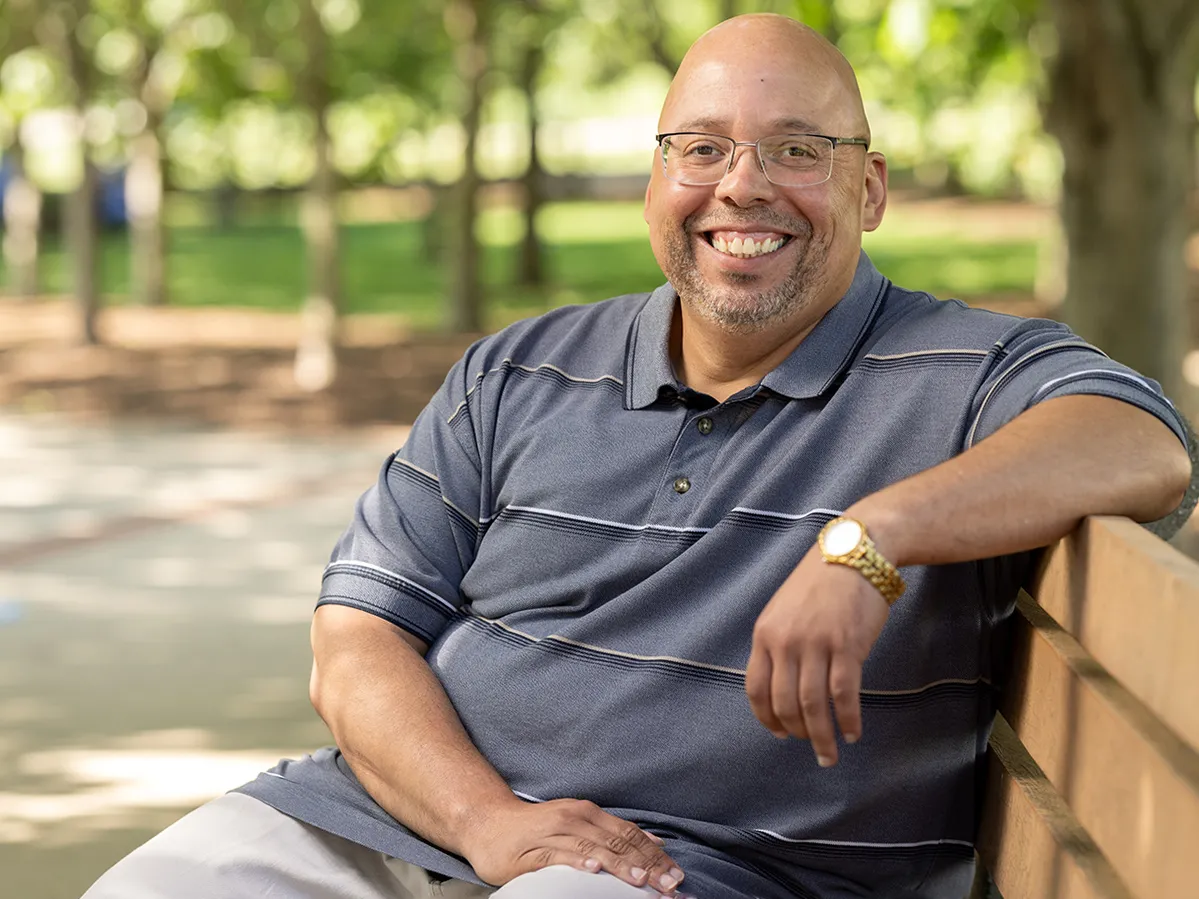 Shawn Lucas, a white man with shaved head and glasses, sits on a park bench with one arm propped along the back. His smile seems both genuine and inviting—he looks like the kind of man students could easily feel at ease with.