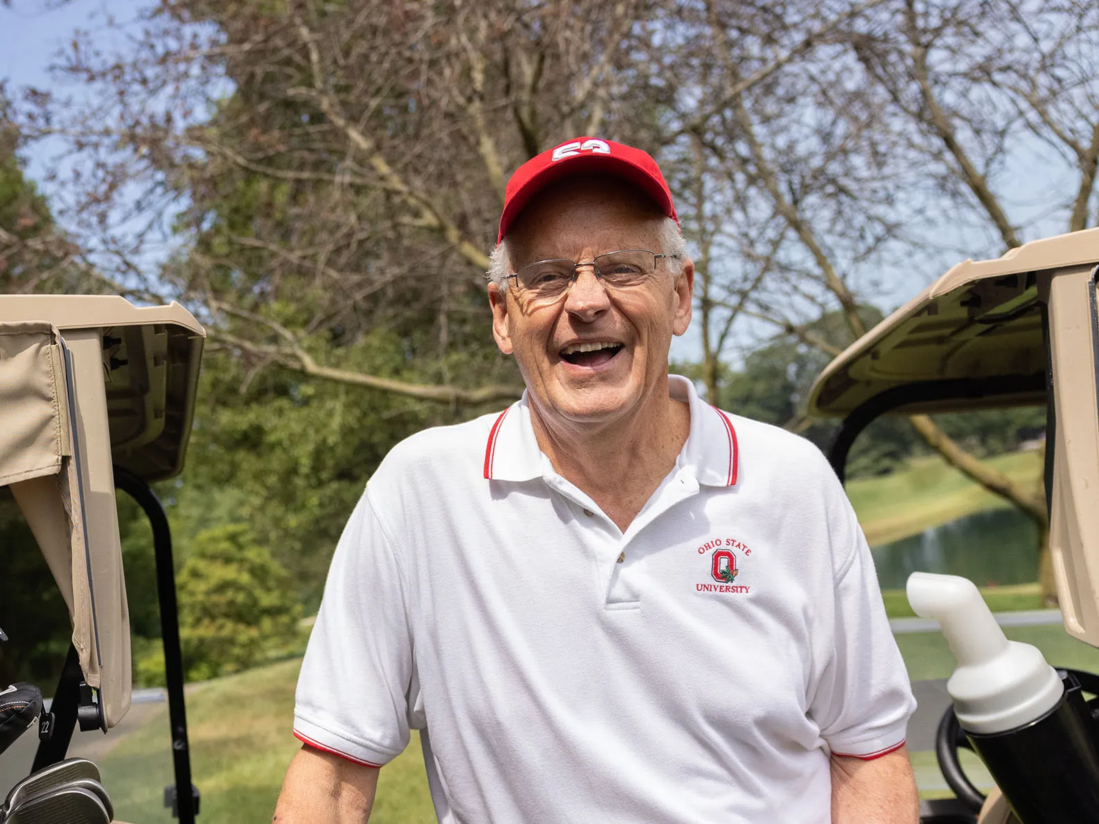 Randy Gradishar, an older white man wearing golfing clothes and a ballcap, slightly leans on a golf cart as he laughs during a conversation with someone not shown in the photo.  