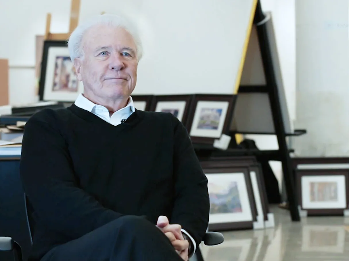 George Acock, an older white man with pure white hair, sits with hands clasped and legs crossed, looking happy, confident and comfortable in a studio where canvasses line the wall and other available space.