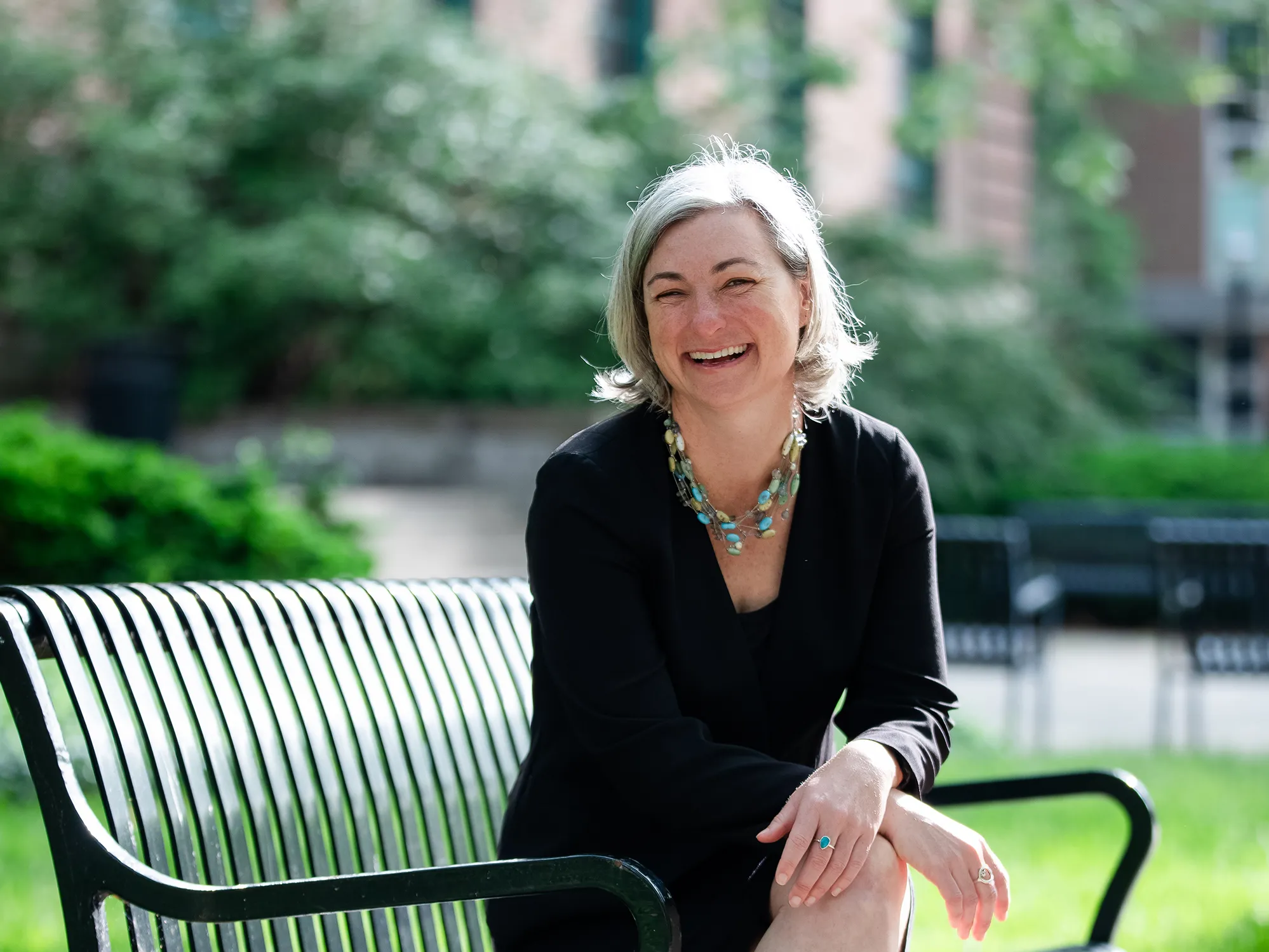 A white woman with shoulder-length gray hair but a young face laughs as she is photographed. Her enjoyment is so true, her eyes are completely squinted. To be fair, it is a bright day on campus. She sits on a metal bench.