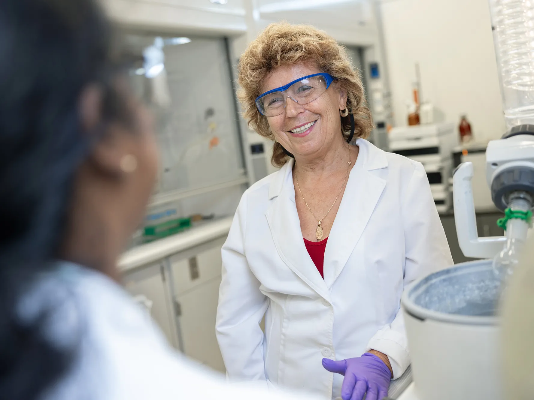 Professor Judit Puskas smiles as she wears a lab coat, safety goggles and talks with a student at a work bench with industrial-seeming equipment. Puskas is a white middle-age woman with blond hair. The student can only be seen from behind. She’s likely Indian or Middle Eastern.