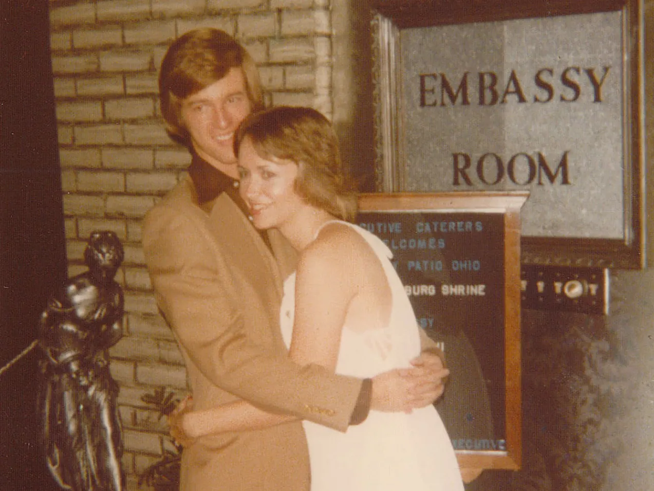 In this old photo, a college-age couple hugs as they pose, clearly comfortable being affectionate with each other. They’re both dressed up and have the feathery hair of the 1970s.