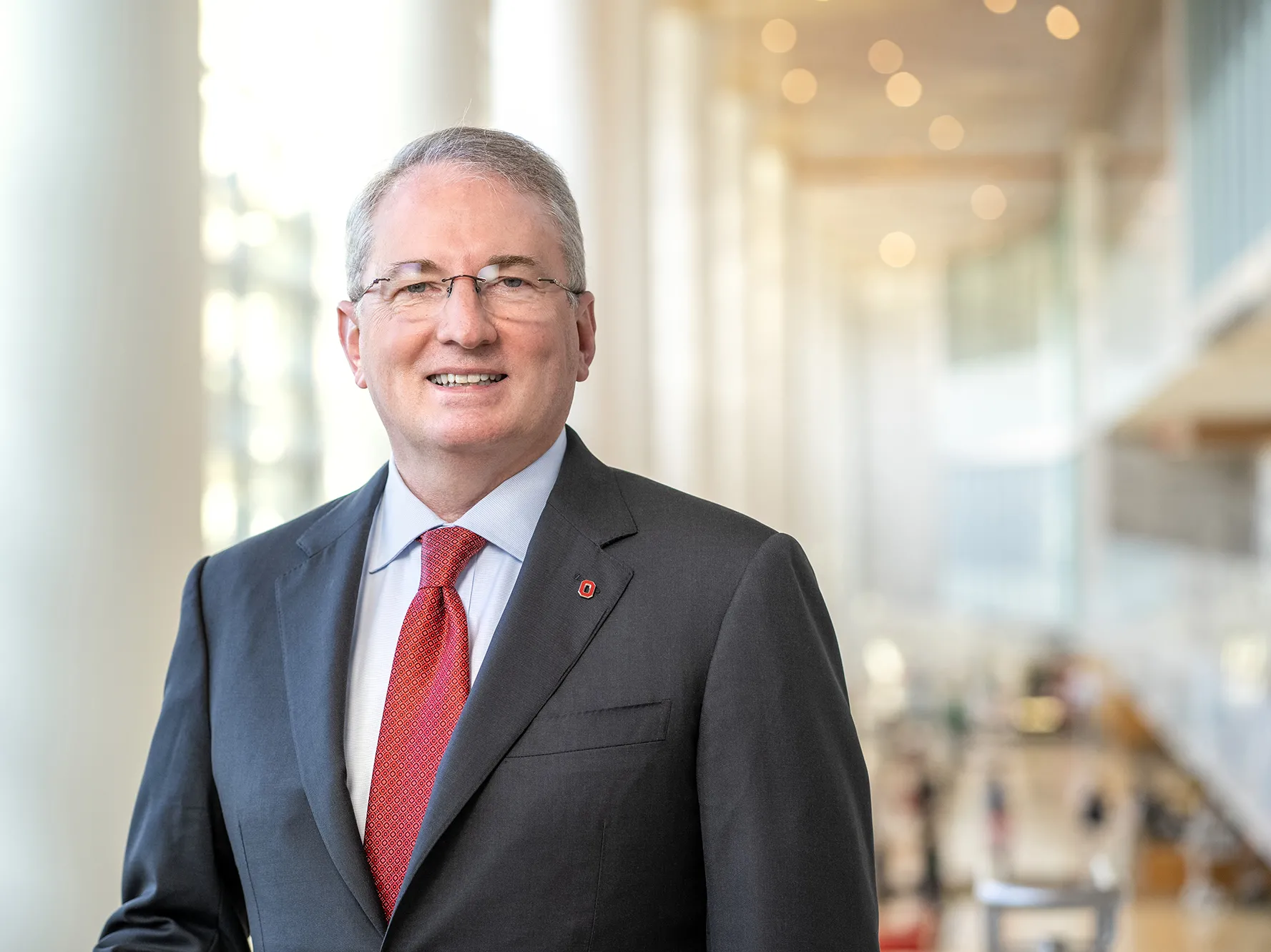 Dr. John J. Warner looks friendly and smart as he poses for a photo in a grand space where pillars and windows line a wall and study tables can be seen behind him. Warner, who is smiling, is a middle age white man in a suit with glasses and a dimple.