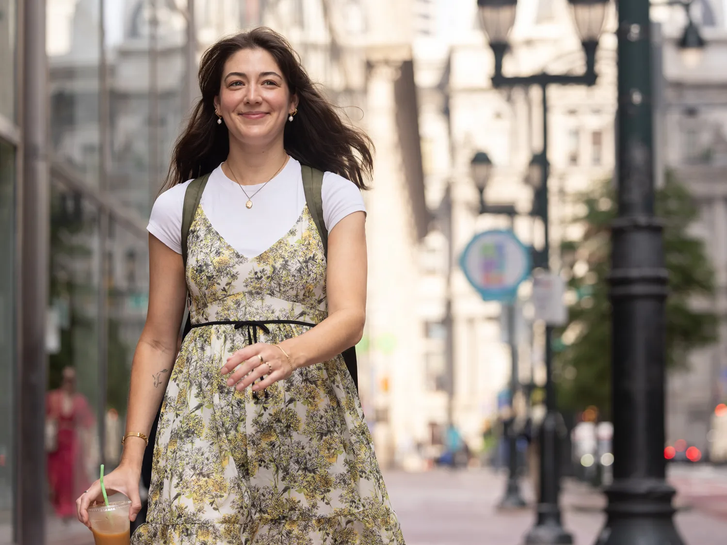 Tovah Kaiser smiles as she strides down a Philadelphia sidewalk. She’s a young white woman with long hair and a tattoo of a simplified fish on the inside of her right arm. She’s wearing a pretty and floaty floral dress over a plain white T-shirt along with gold earrings, necklace, bracelet and rings. 