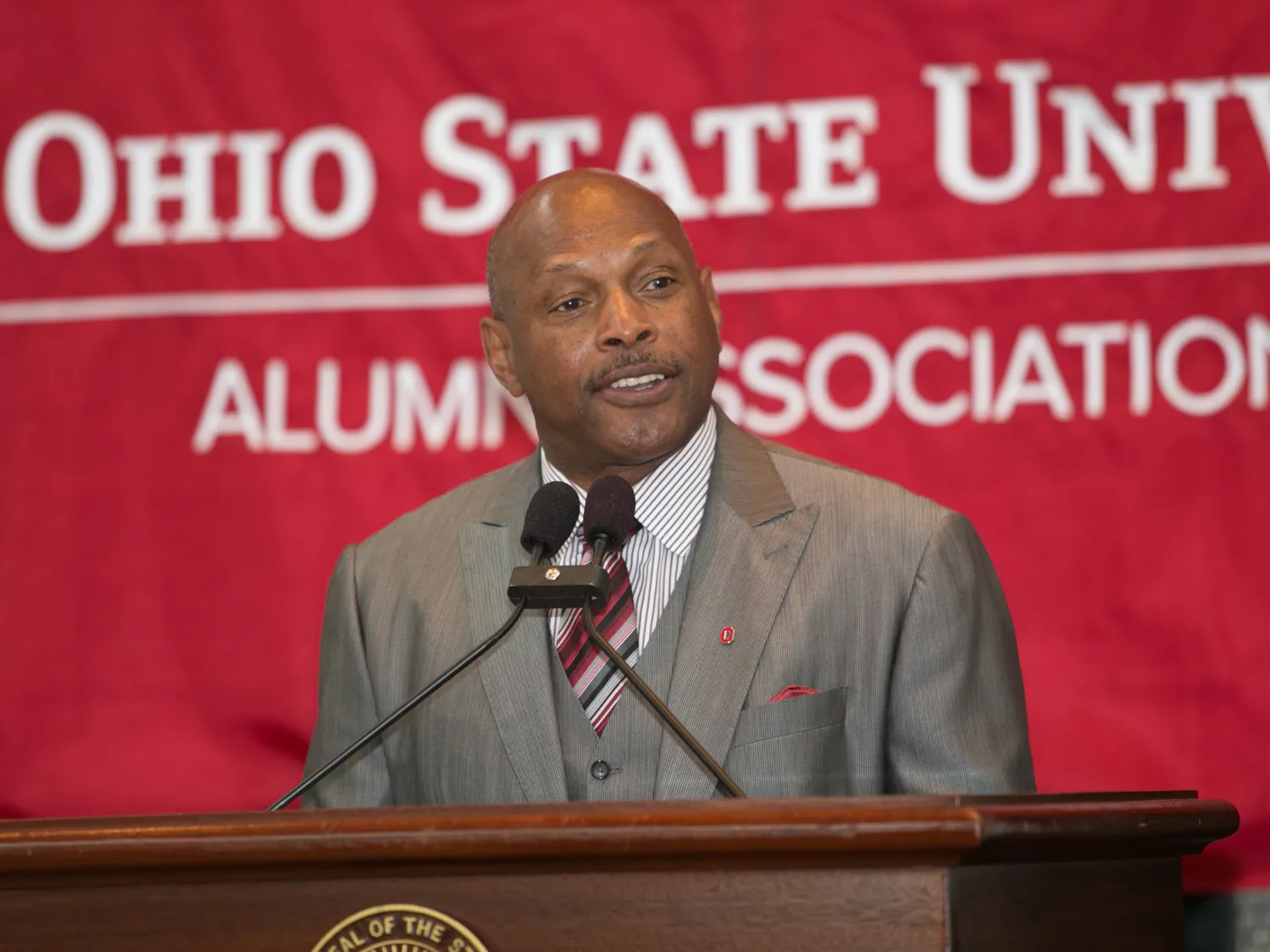 In a three-piece suit, Griffin stands behind a lectern with two microphones. Behind him is a giant banner that says “Ohio State University Alumni Association.” He’s smiling while he listens to someone off camera talking or asking a question.