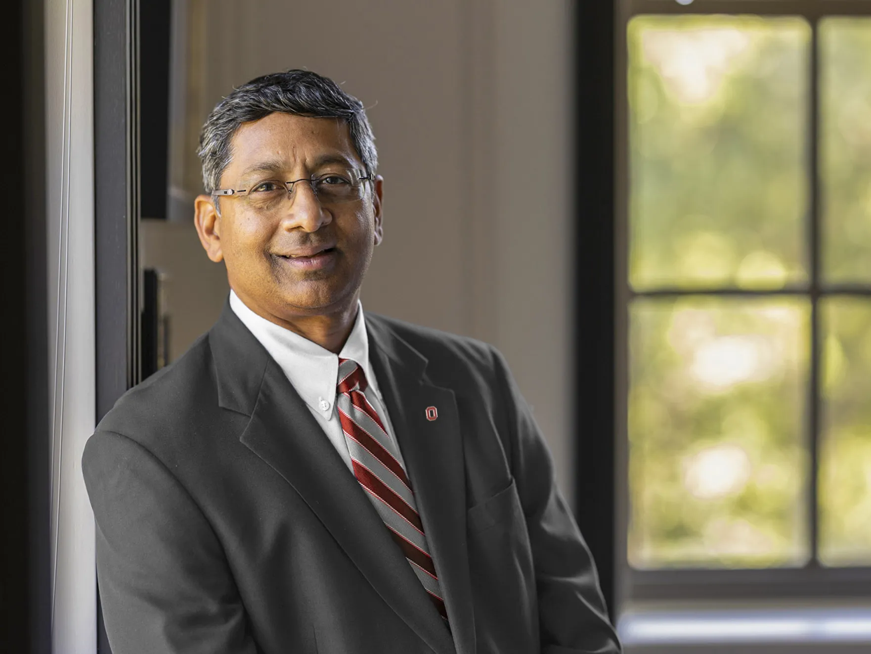 Ravi V. Bellamkonda looks calm and capable as he smiles for a photo in an academic building with a window out of focus behind him. Ravi, a middle-aged Indian-American man, wears wire-rim glasses and a suit.