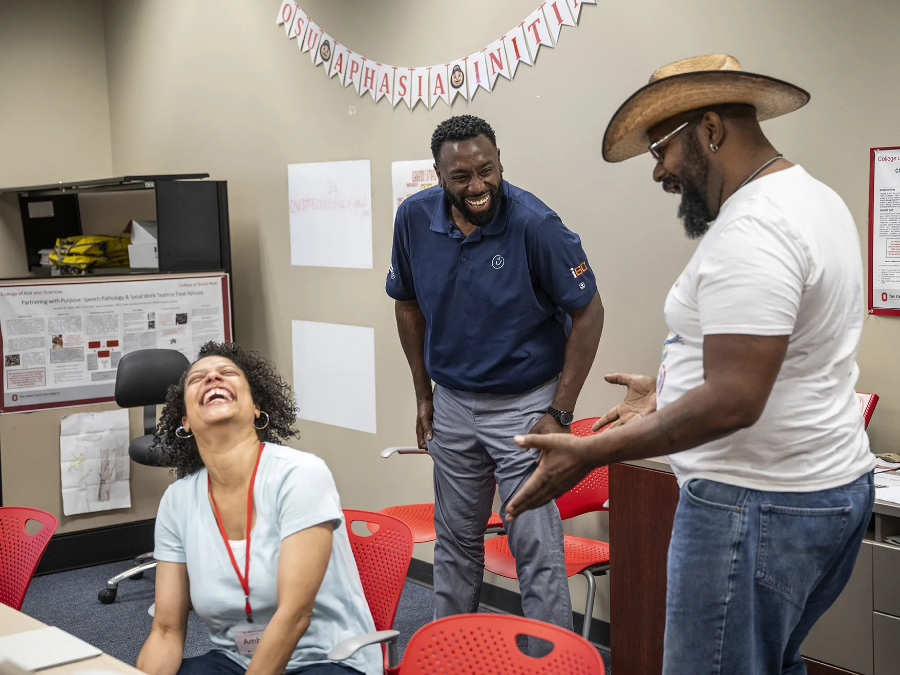 Three people laugh as they talk in a room with flags spelling OSU Aphasia Initiative on the wall. Two of them are husband and wife. She has her head thrown back as she laughs hard; her hoop earrings swing with her movement. Her husband, in dress pants and golf shirt, laughs with his hands on his thighs as he watches the man entertaining them with his storytelling. That man wears a cowboy hat and glasses. All three are black and middle-aged.