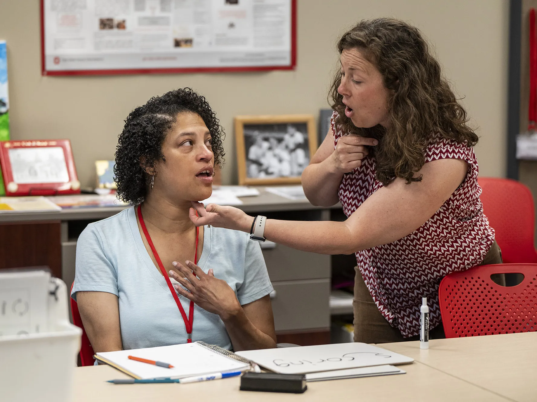 Amber Poindexter, sitting at a table, touches her chest as she watches the mouth of her speech therapist, Arin Sheeler. Arin is a curly-haired white woman leaning toward Amber, pointing at her own throat and touching the underside of Amber’s chin. They’re very focused on each other.