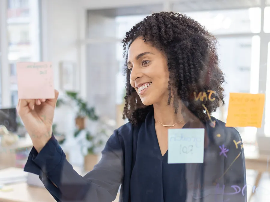 A young woman of mixed race smiles as she writes on a sticky note attached to a see-through project space in an office setting. The notes could be used for planning or leaving encouraging messages for co-workers; the text can&#039;t be read in the photo.