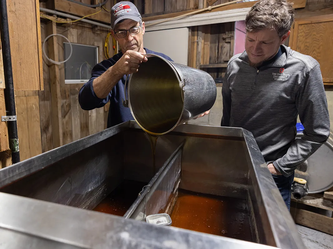 One man holds a bucket as he pours cooked sap into one side of a stainless steel maple-syrup boiling stove. Another man, Karnes, watches. They're inside a rustic looking shed on Brown's property.