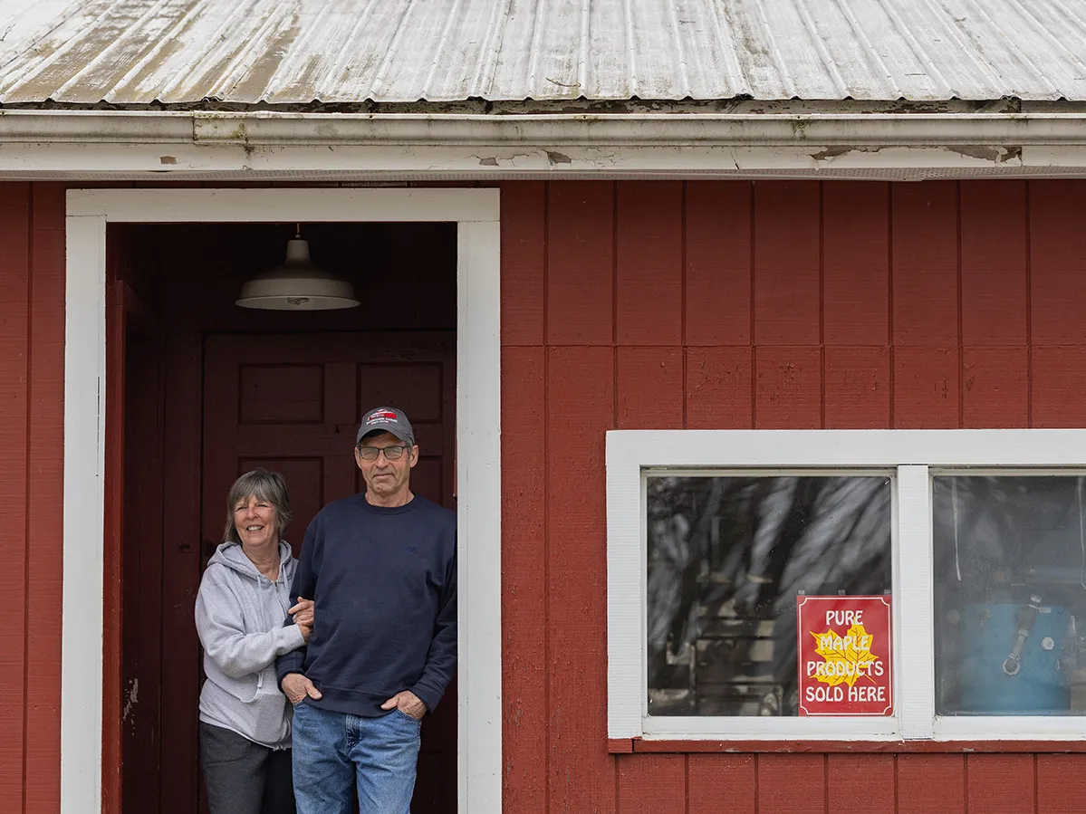 A husband and wife smile as they pose for a portrait in the doorway of of a building they use to cook maple syrup on their property. The building has a classic fall red-and-white design and is picturesque.