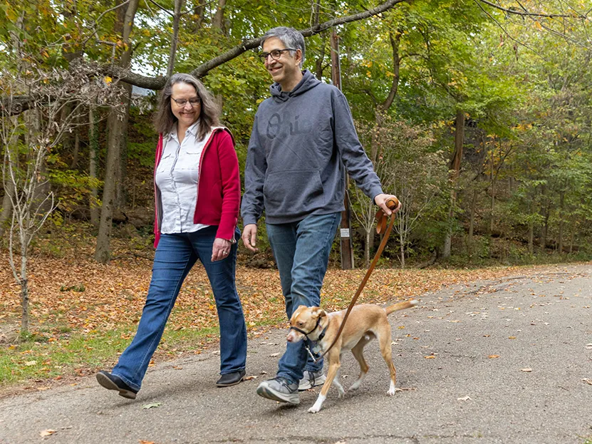 A smiling couple walks their dog through woods on a paved path