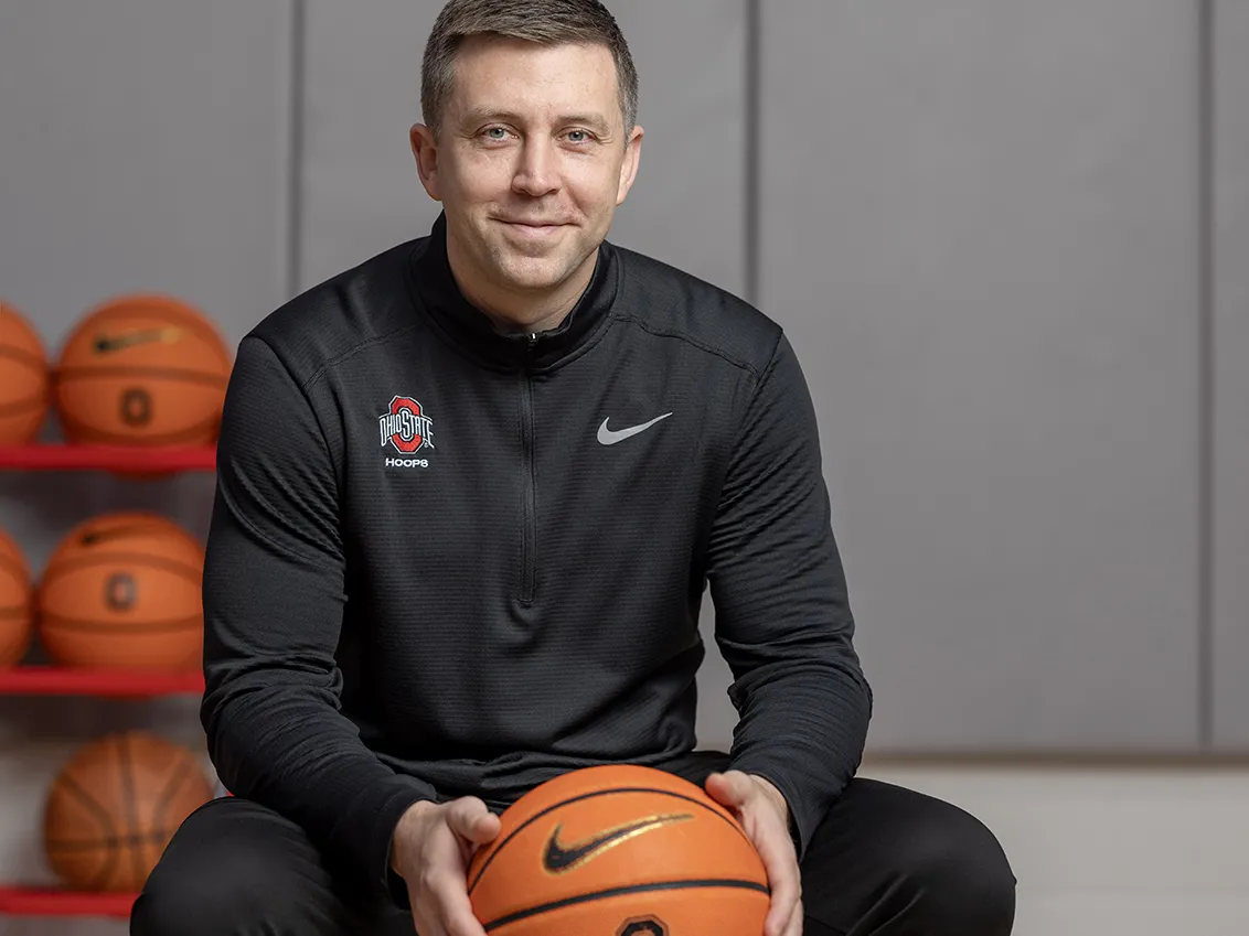 Ohio State Basketball Coach Jake Diebler holds a basketball while sitting on a stool and smiling warmly at the camera. He looks like he is about to laugh. Diebler is a white man who looks to be in his 30s. He has short cropped hair, blue eyes and wears black workout gear.