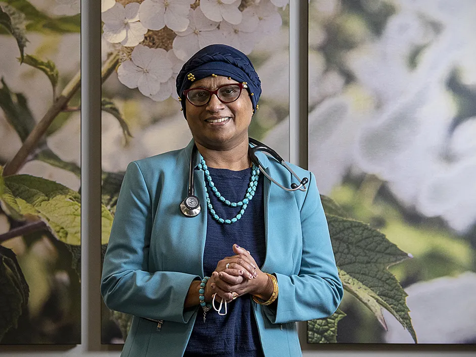 Dr. Bhuvaneswari Ramaswamy wears a headscarf wrapped around her head and glasses as she smiles for a portrait. At this time, she was undergoing cancer treatment and caring for patients. She looks liek someone you'd want to trust. A middle-age woman born in India, she wears a business suit, a stethoscope around her neck and turquoise necklace and bracelet. The background is a wall with a huge photograph of flowers.