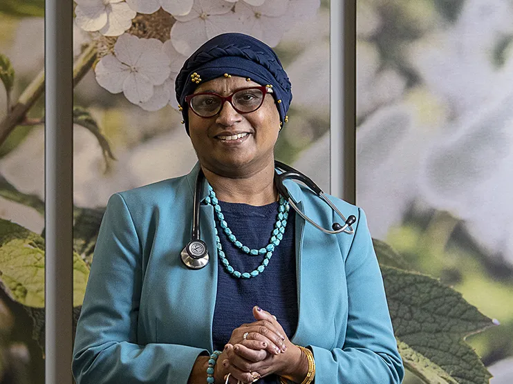 Dr. Bhuvaneswari Ramaswamy wears a headscarf wrapped around her head and glasses as she smiles for a portrait. At this time, she was undergoing cancer treatment and caring for patients. She looks liek someone you'd want to trust. A middle-age woman born in India, she wears a business suit, a stethoscope around her neck and turquoise necklace and bracelet. The background is a wall with a huge photograph of flowers.