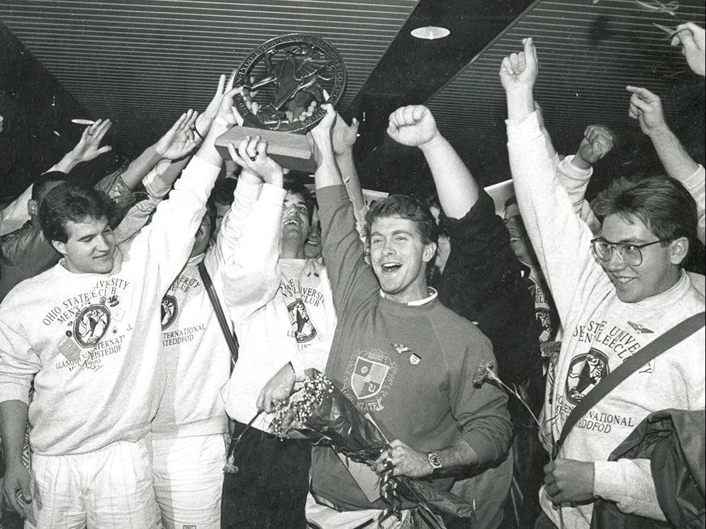 About a dozen members of the men&#039;s glee club hoist a trophy as they cheer. The photo is black and white, giving it a dtaed look, even though color photography was available at the time.