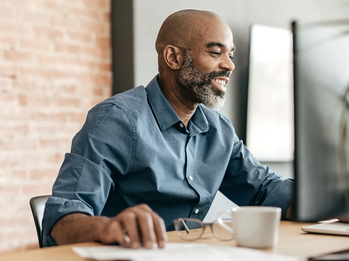 This image shows a Black man with a bright smile sitting at a desk and using a computer