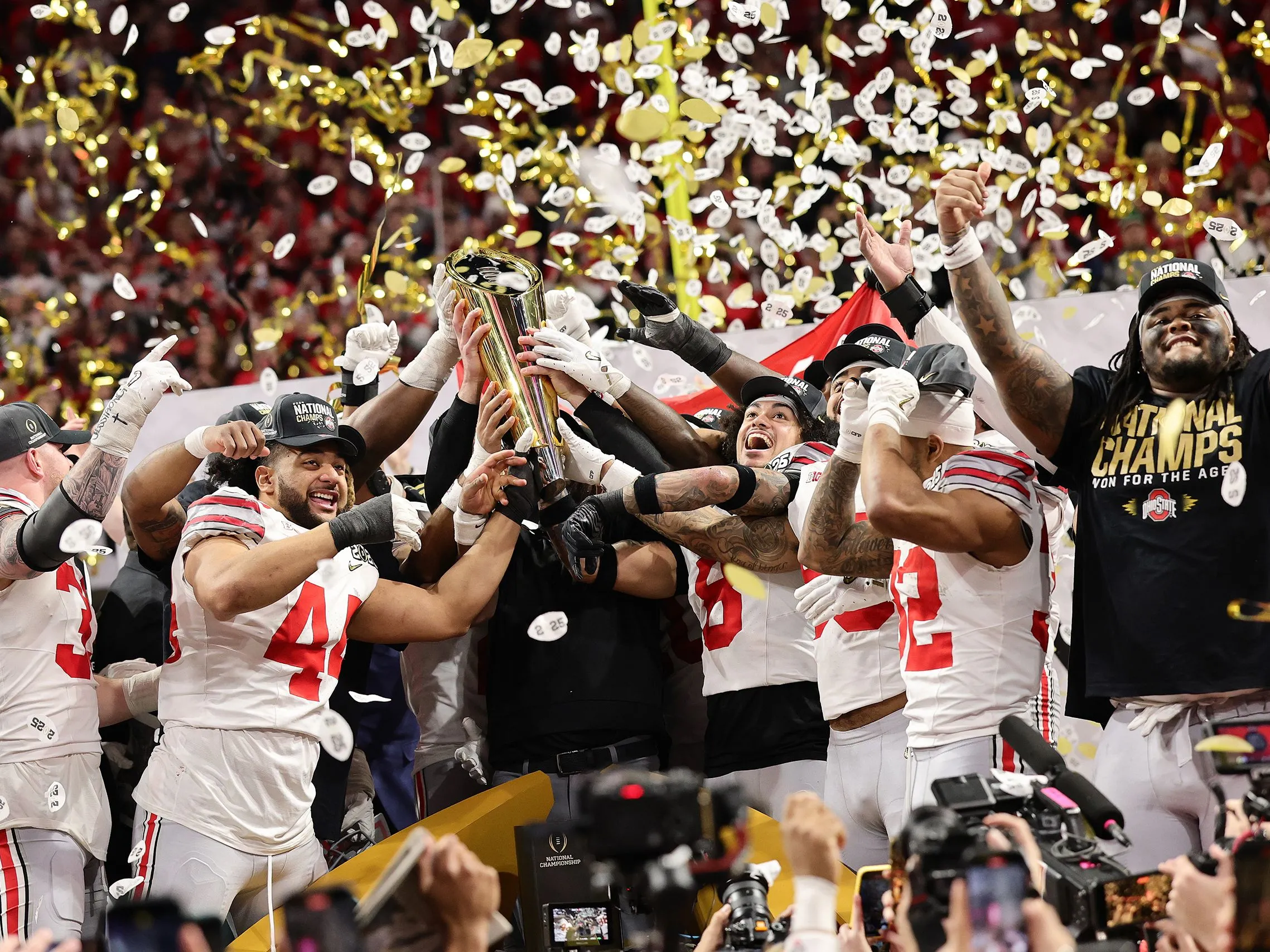 Buckeye football players cheer as they hoist up the national championship trophy. Confetti falls and they&#039;re amidst a huge crowd.