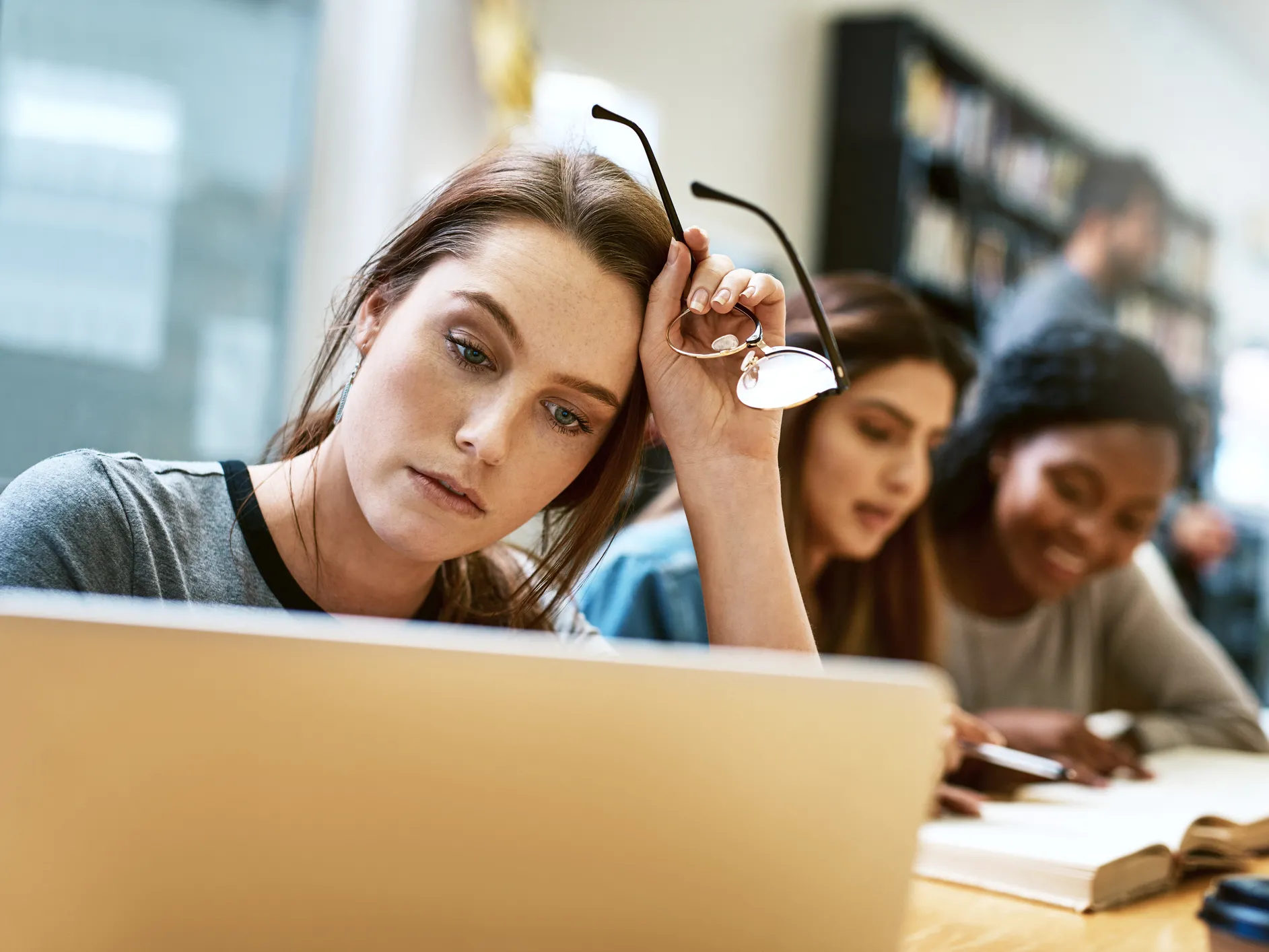 A stressed-out college student has her glasses off but looks at her computer screen while others in the background happily study.