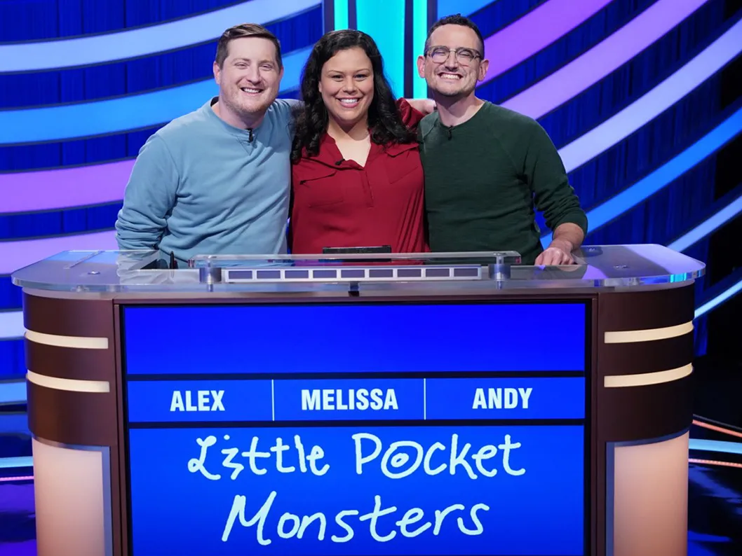 Three Buckeyes pose with their arms around one another on the stage of Jeopardy! The two on the sides are men; in the center is the woman. They seem genuinely close and pleased to be competing together.