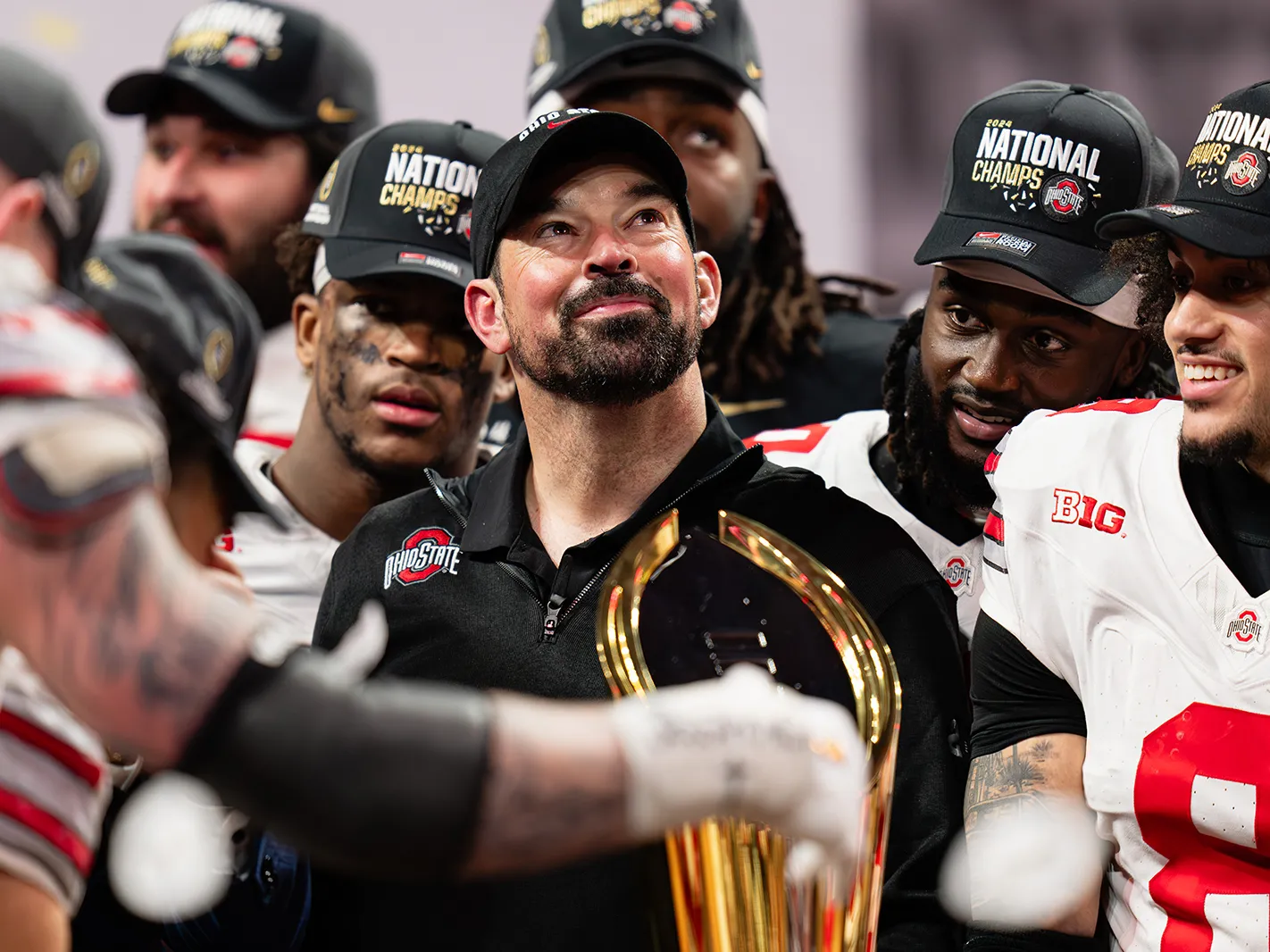 Ohio State football coach Ryan Day, surrounded by football players, looks proudly upward. He appears a little tearful but is smiling. Everyone in the photo taken after they won their final game of the playoffs wears a National Champs baseball cap.