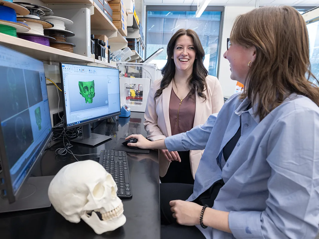 Two women talk in front on a computer monitor showing a 3-D skull. The 3-D printed result is on the table next to them.
