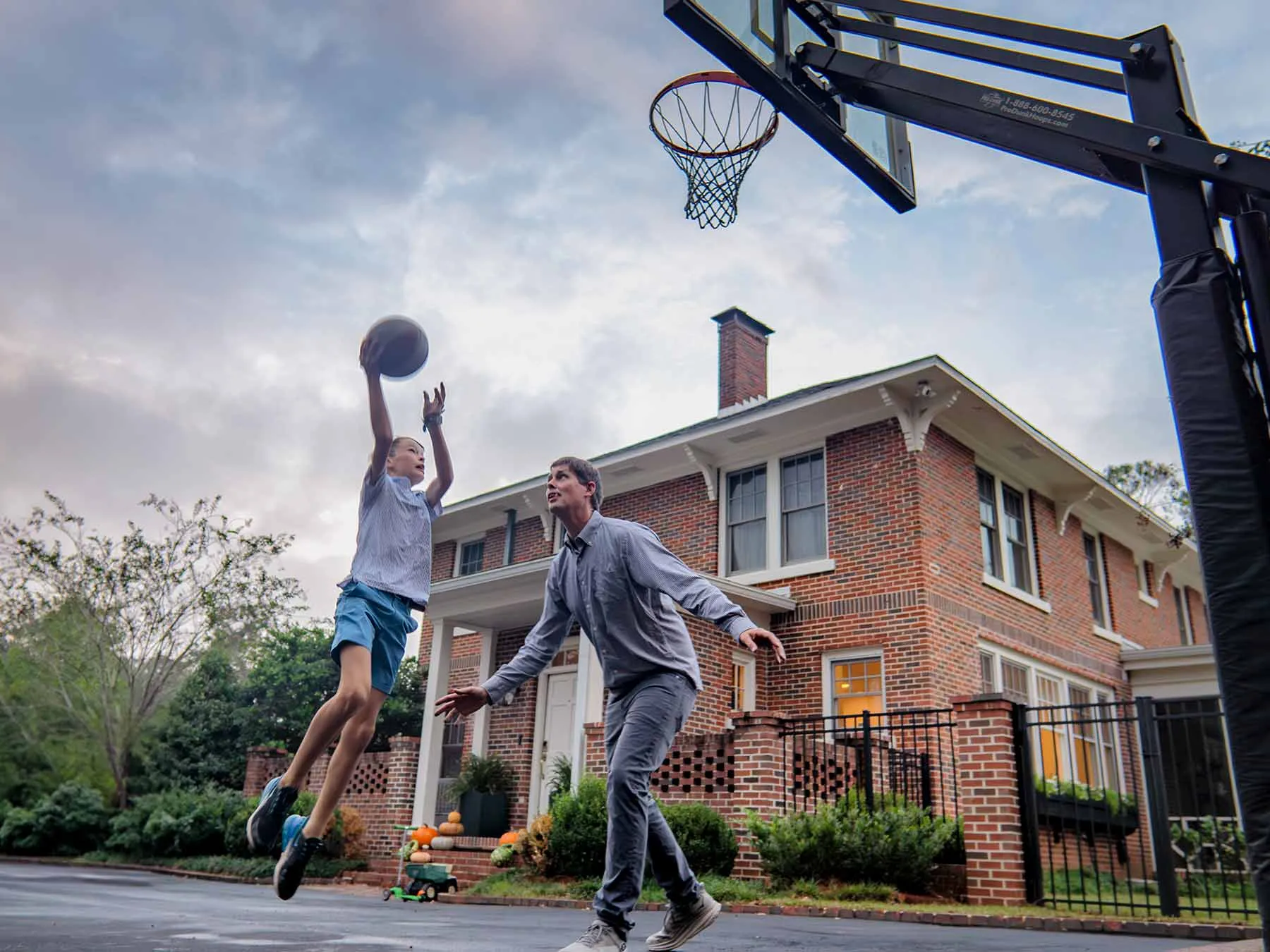 10-year-old Byron jumps as he shoots the basketball toward the net in a pickup game of basketball in the driveway at his home. He&#039;s a lanky white boy in gym clothes. His dad wears jeans and a button-up and they are intensely focused. Their brick two-story home is in the background.