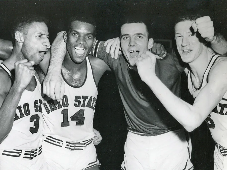 Four basketball players and their coach grin as they pump their fists in celebration after winning a big game. Two of the players and the coach are white and two of the players are black. They all have that clean cut air of the late 1950s and early 1960s.