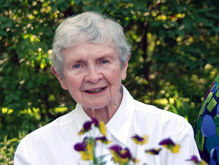 Sister Mary Faith Dargan, an older white lady, smiles as she poses for a portrait outdoors. She wears a plain white button-down that makes her blue eyes and smile stand out