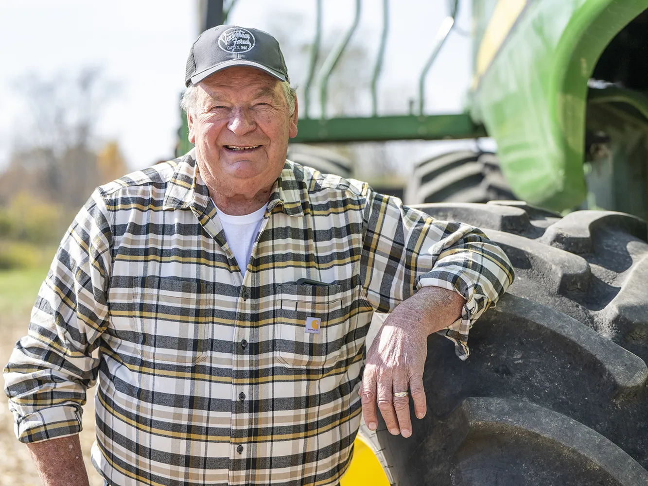 Len Vonderhaar is an older white man wearing a plaid shirt and smiling happily as he leans on a wheel of his tractor