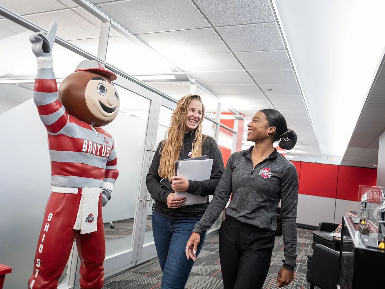 A statue of Brutus Buckeye waves his No. 1 finger as a young white woman and a young black woman walk by happily chatting.