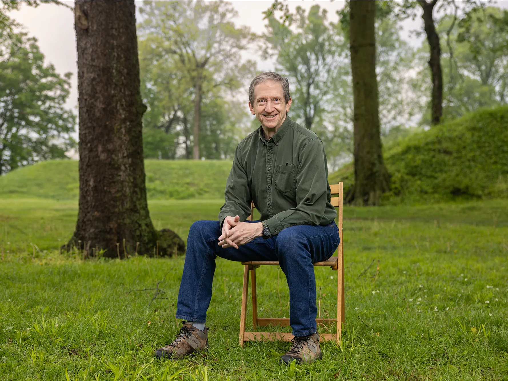 Bradley Lepper, a gray-haired white man in jeans and an earthy colored button down, sits on a chair at the Great Circle Earthworks. Around him are trees and behind him, grass-covered mounds rise. The portions of the mounds in the photo look to be about as tall as an adult, or at least shoulder height.